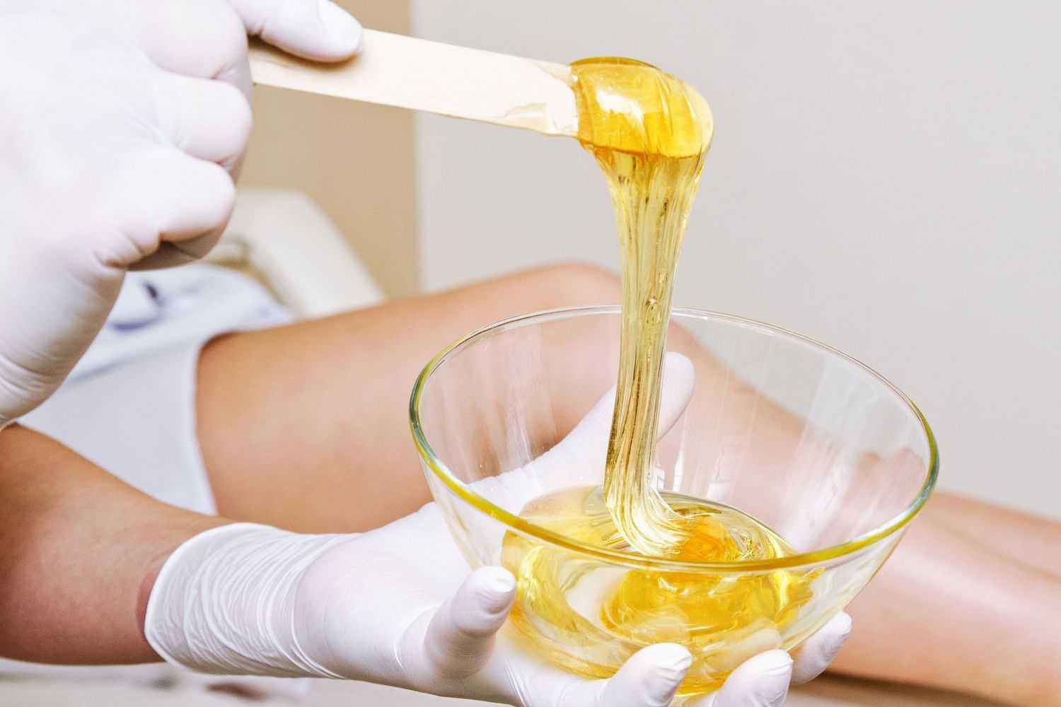 Gloved hands lifting a wooden spatula with dripping honey wax.